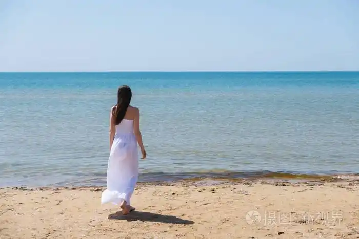 年轻的黑发女人,在夏天白色礼服站在海滩上,望海