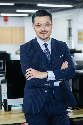 身着蓝色西装三件套的男士靠坐在办公室的桌子上自信双手抱拳成功人士