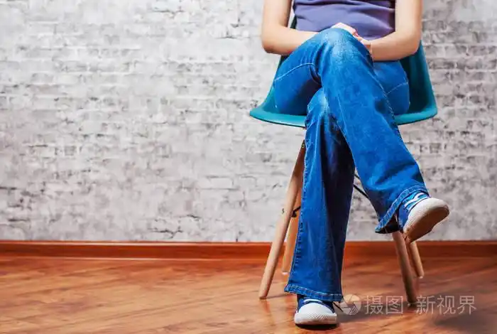 穿牛仔裤和运动鞋的女性双腿坐在灰色墙壁背景的椅子上,有复印空间