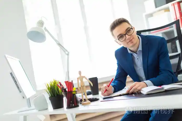照片 一个年轻人坐在办公室的一张桌子旁, 手里拿着一支红色的钢笔