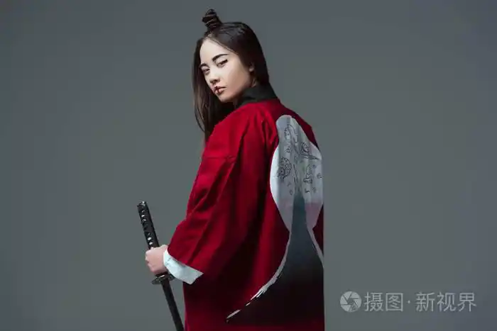 年轻女子在传统的日本和服手持武士刀, 看着相机孤立灰色