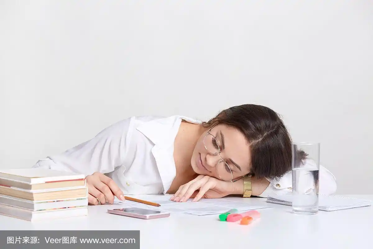 过度劳累的黑发学生斜靠在桌子上,在工作地点打盹,感到疲倦,穿着白