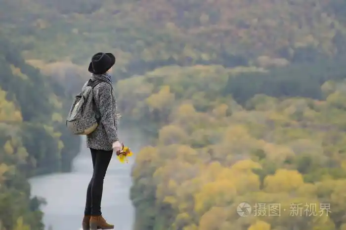 一个背着背包,戴着帽子站在山上的女孩.举起手来.下面是河流和山脉.
