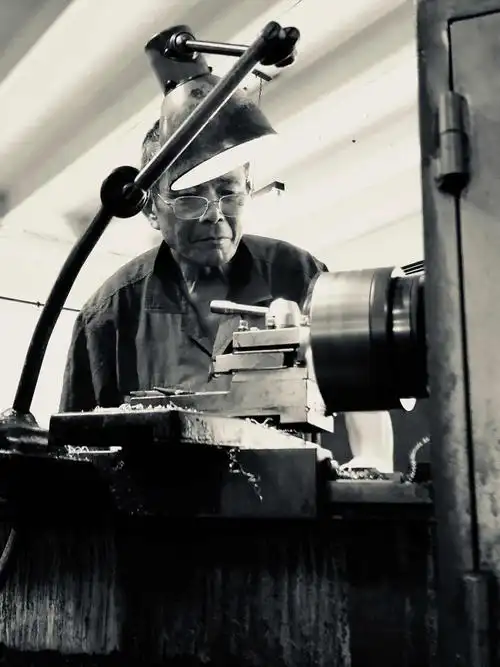 一个老工人一丝不苟的在台灯下的金属机器前做事 整个车间里也只有他
