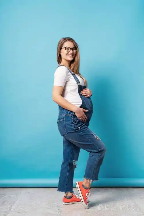 一个可爱的年轻女人戴着眼镜和牛仔裤,摆着摸肚子,与她未出生的孩子
