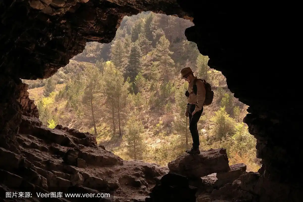 登山探险家戴着帽子和背包进入一个山洞俯瞰一个美丽的山谷与绿树在