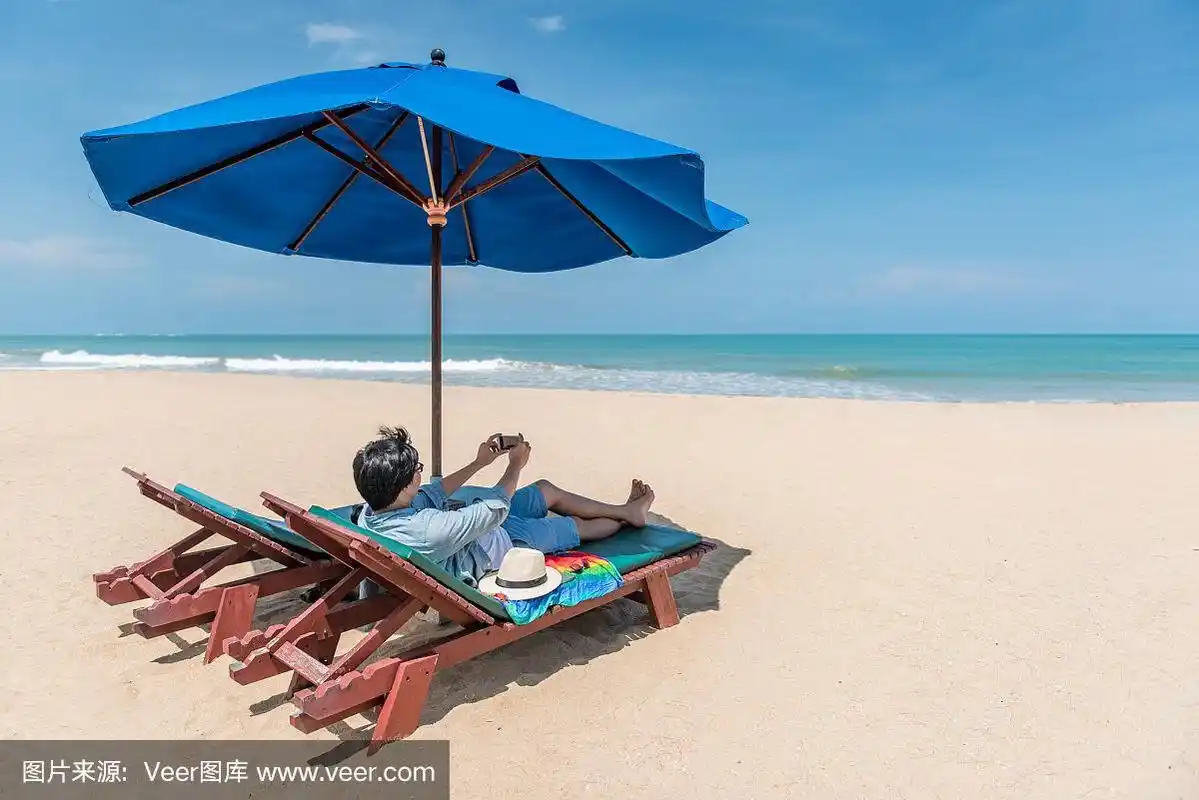 一个年轻人躺在沙滩的木制长椅上,用智能手机拍照