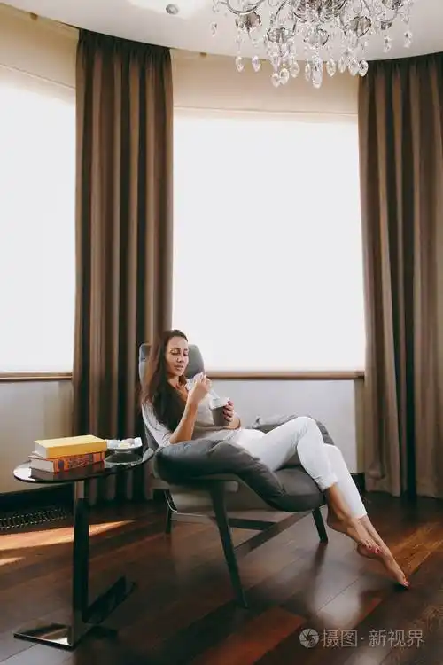 美丽的年轻女子在家坐在现代的椅子上在窗前在她的客厅里放松喝咖啡或