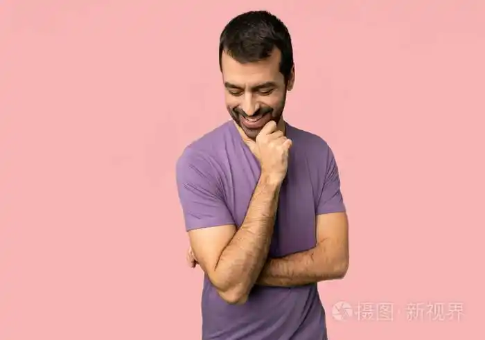 英俊的男人低头看着,手放在孤立的粉红色背景上的下巴上