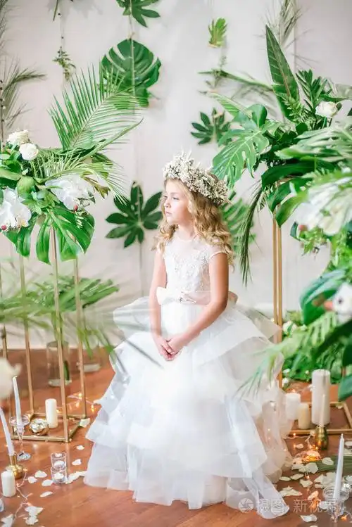 照片 可爱的小女孩,一头金发,身穿白色婚纱,戴着热带树叶,白玫瑰和