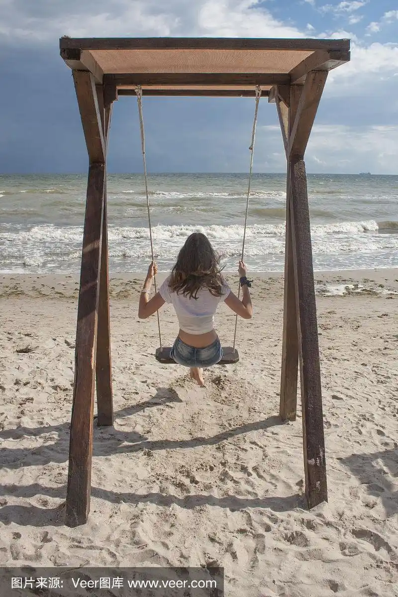 穿着牛仔短裤和白色t恤的年轻女子在海边的秋千上荡秋千.