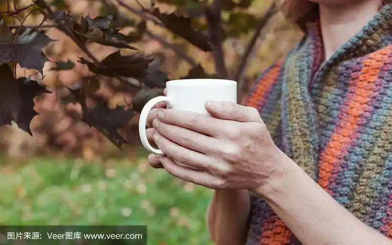 优雅的女性双手握着一个白色的杯子,里面盛着热饮.秋天的背景.