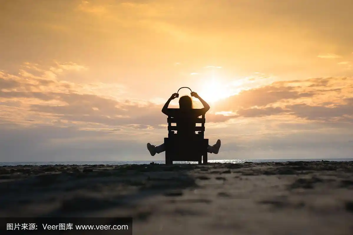 戴着耳机的女孩坐在躺椅上,在日落时分的海滩上听着音乐.夏天的概念.