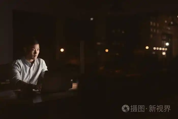 照片 一位年轻的亚洲商人深夜独自在办公室的办公桌前加班,背景是城市